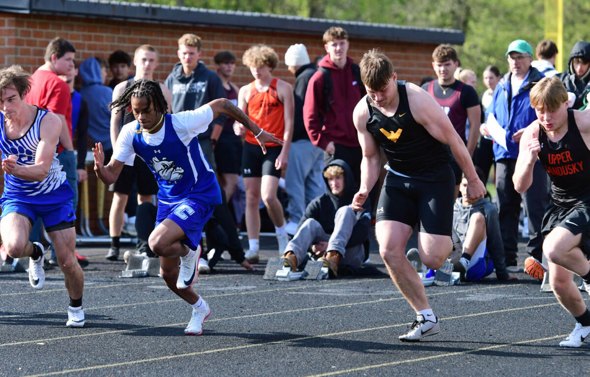 Four male sprinters race off the blocks on a track as a crowd watches nearby near a brick wall.