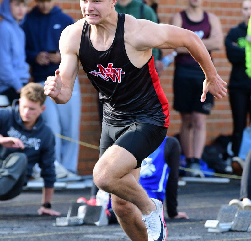 Male sprinter in a black and red uniform mid-stride on a track, determined expression, with spectators in the background.