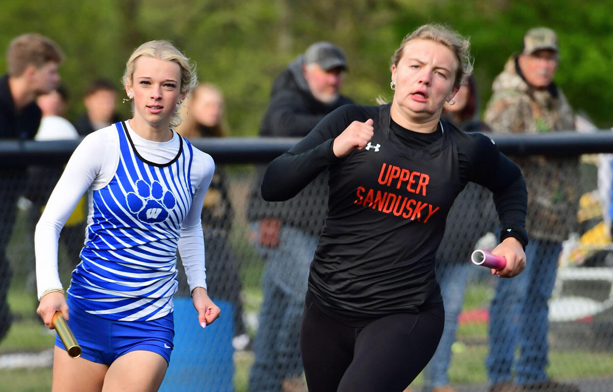 Two female relay runners sprint on a track: one in a blue and white striped uniform, the other in a black 'Upper Sandusky' jersey, each holding a baton and vying for position.
