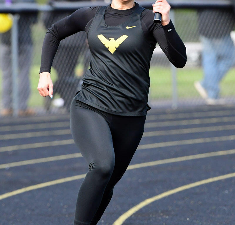 Female sprinter running on a track while holding a baton, wearing a black kit with a gold WV logo and pink shoes.