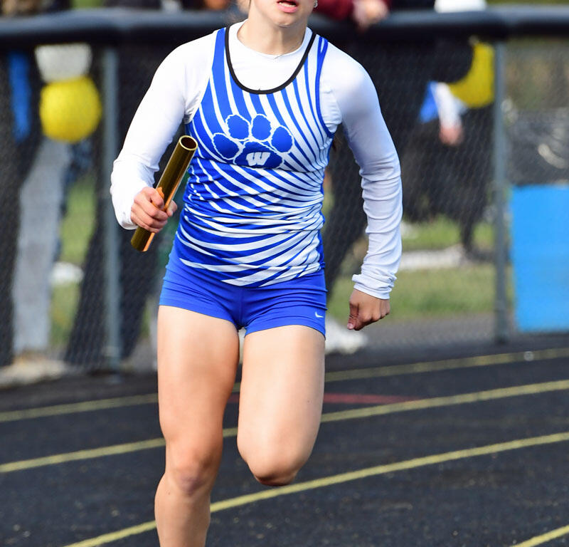 Female athlete running with a baton in a relay race on a track, wearing a blue and white uniform with pink shoes.