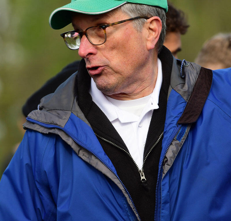 Man wearing a green cap, glasses, and a blue jacket outdoors, speaking to someone off-camera.