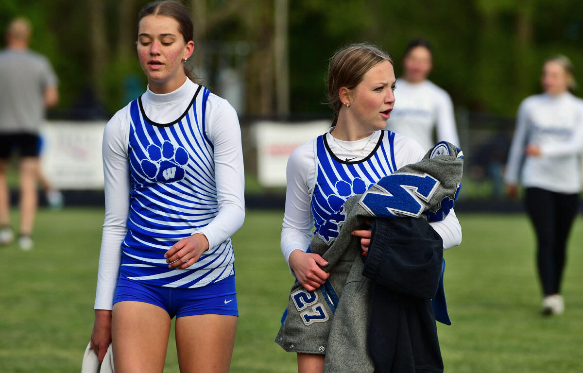 Two female track athletes in blue-and-white striped uniforms walk on a grassy field, one carrying a gray jacket.