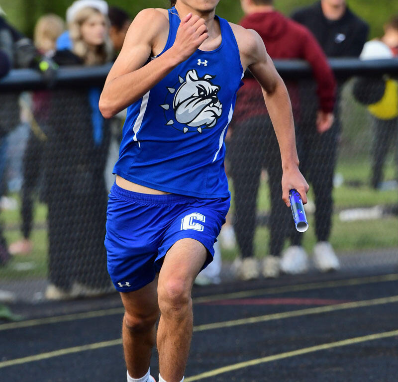 Male sprinter in a blue jersey with a bulldog logo, running on a track while holding a baton, with spectators in the background.