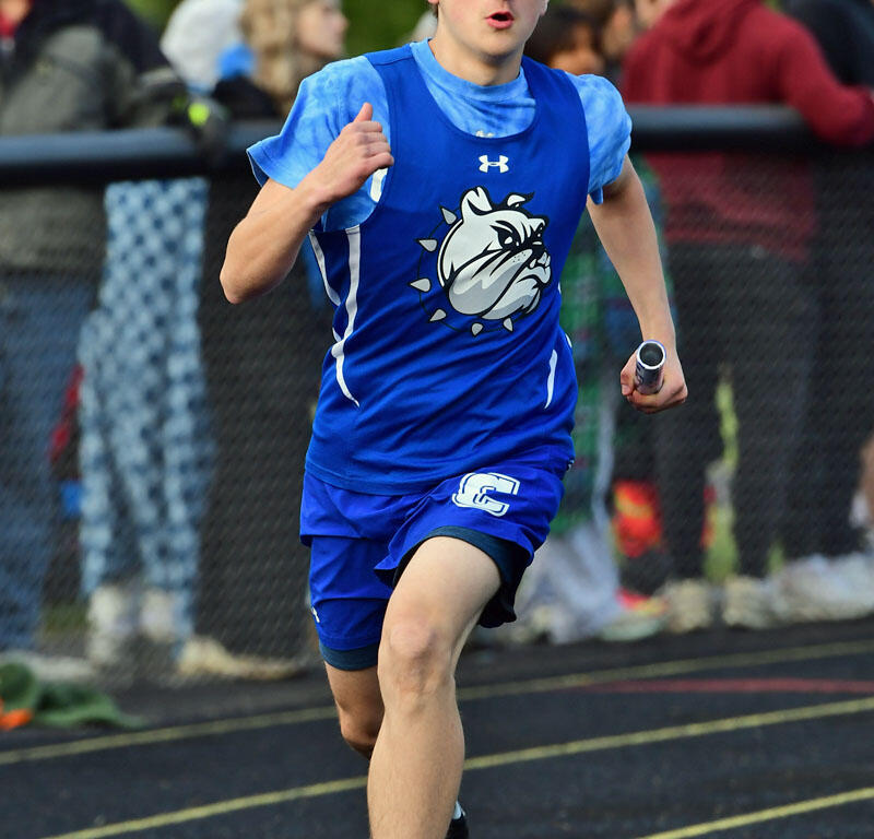 Young male runner in a blue jersey sprinting on the track as spectators watch from the sidelines, his expression focused.