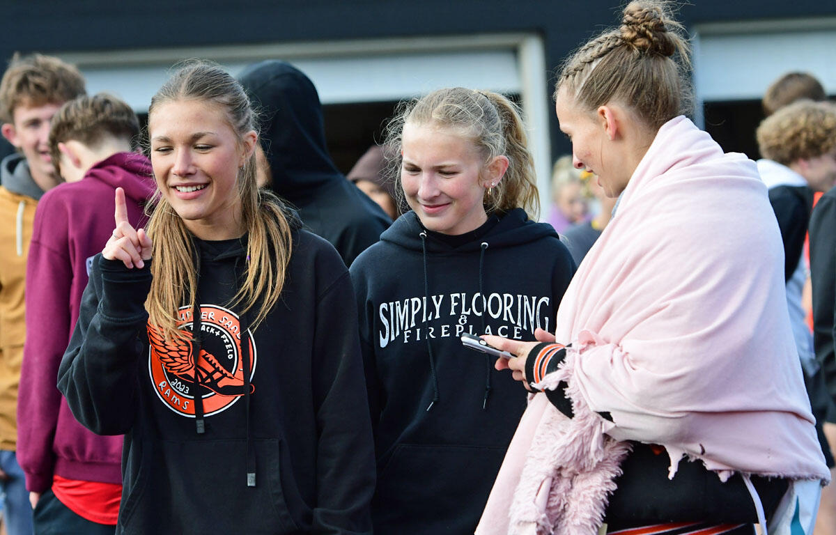 Three teenage girls in hoodies smile and chat outdoors, with one girl raising a finger in greeting or emphasis.