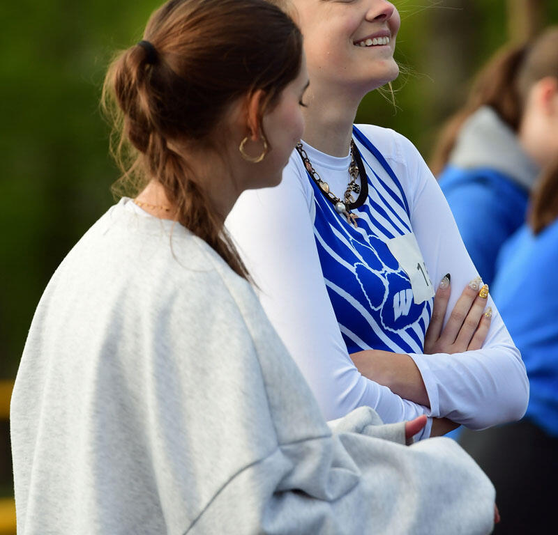 Two women stand outdoors, smiling and chatting; one in a gray hoodie and the other in blue-and-white athletic wear.