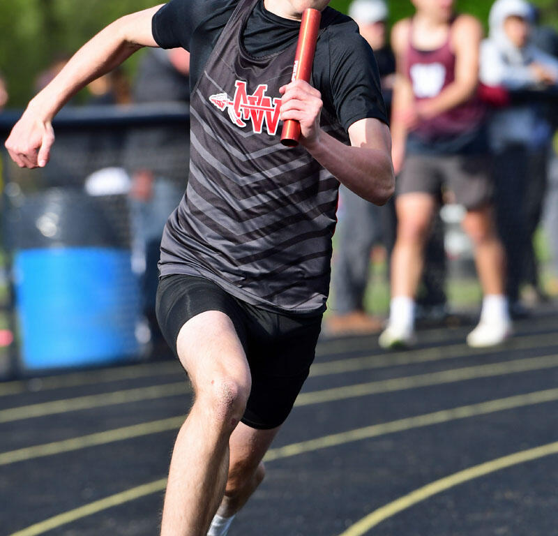 Male sprinter in a black-and-gray uniform runs with a red baton during a relay on an outdoor track, with spectators in the background.