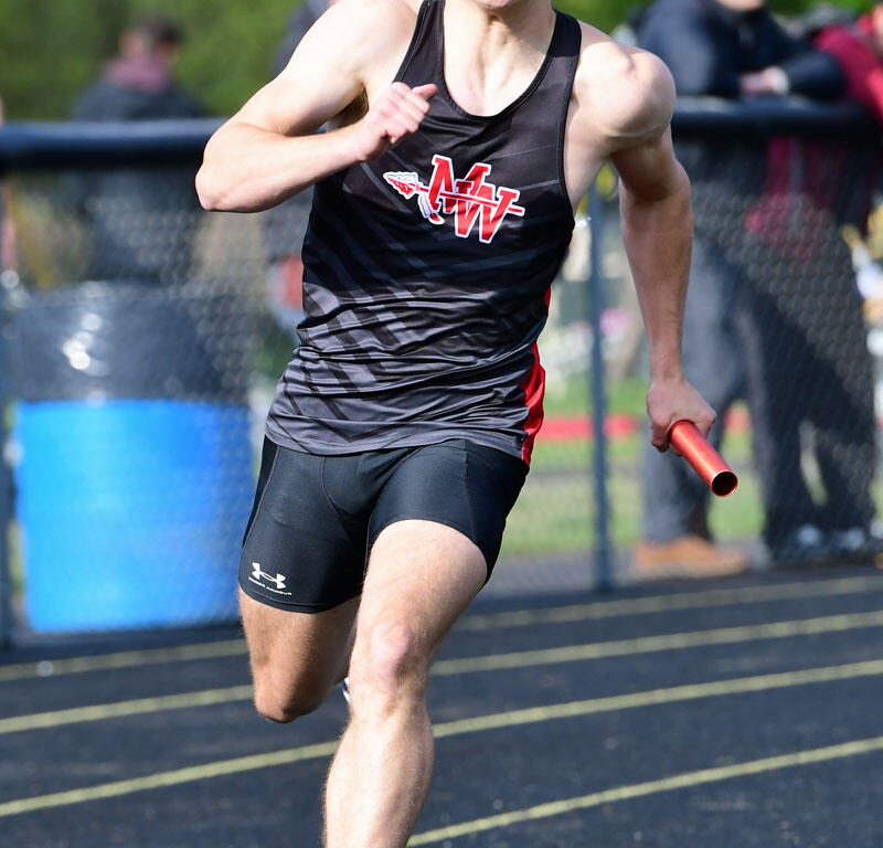 Male sprinter in black and red uniform running on a track, holding a red baton in a relay race presence of spectators behind a fence.