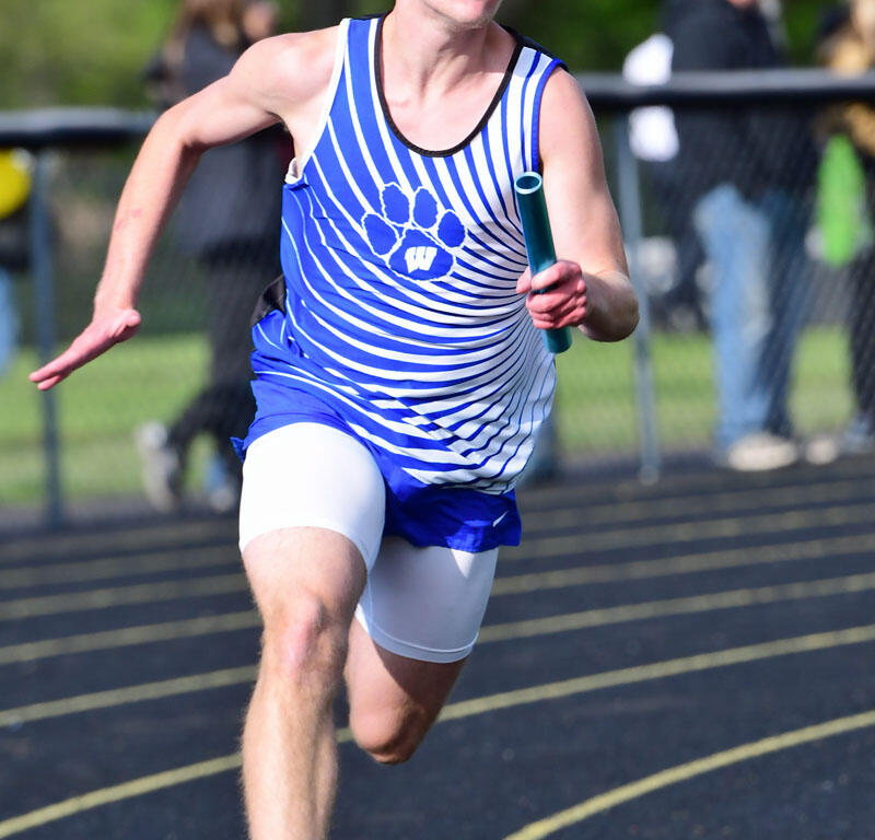 Young male sprinter in a blue and white striped jersey holding a baton, running on an outdoor track.