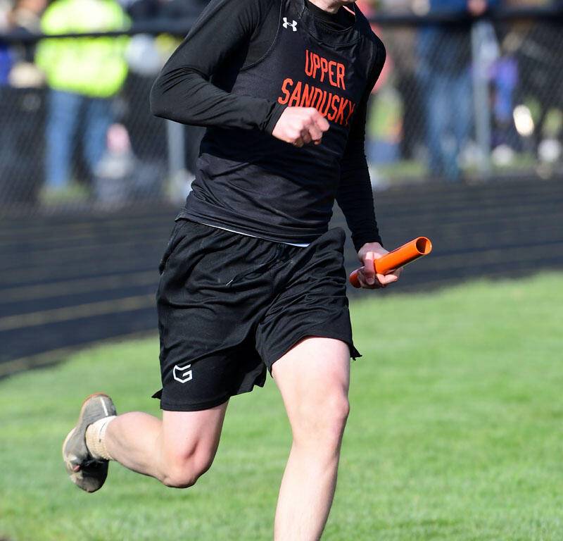 Sprinter wearing a black Upper Sandusky uniform runs on a track, holding an orange baton as spectators watch in the background.