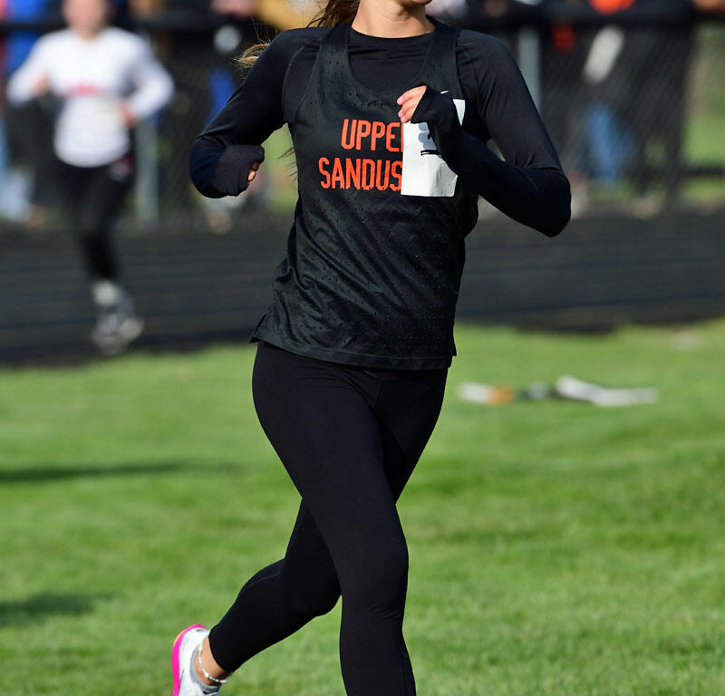 Female runner mid-stride on a track, wearing black running gear and a bib number, with spectators in the background