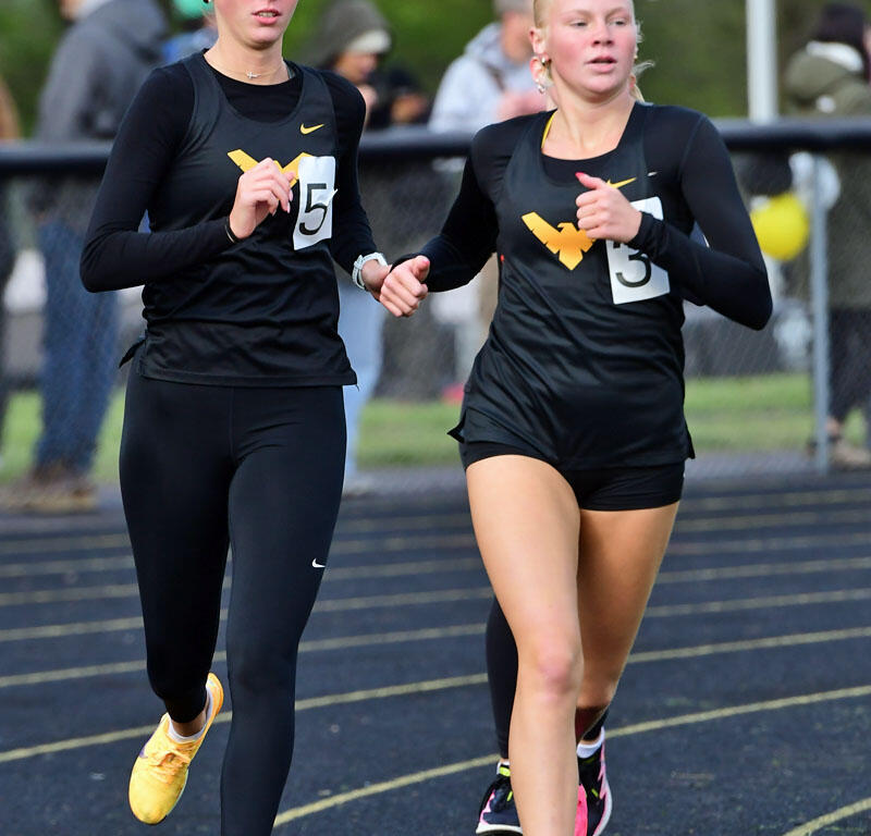 Two female runners in black outfits hold hands while sprinting on a track during a race, wearing bib numbers 5 and 3.