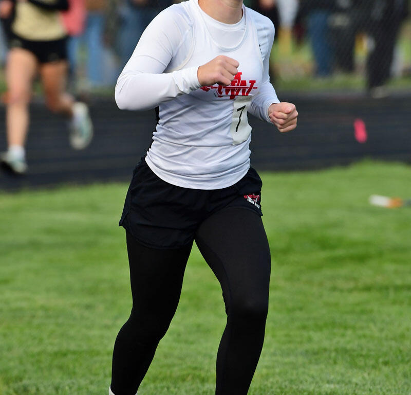 Female runner on a track mid-race, wearing a white long-sleeve top and black tights, bib number 7, with spectators in the background.