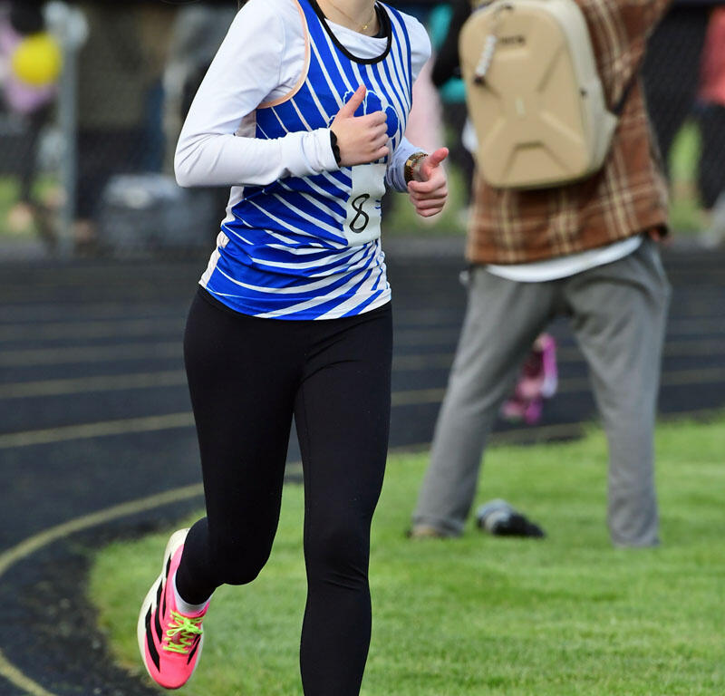 Female athlete running on a track in a blue and white striped jersey, black leggings, and neon pink sneakers.