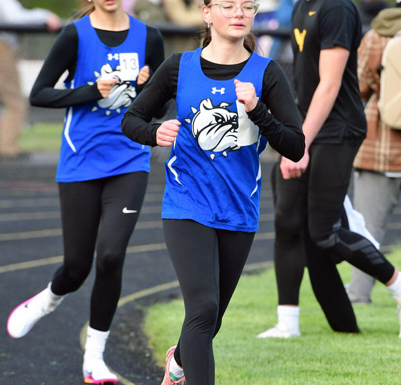 Two female runners in blue jerseys sprint on a track, number bib visible for one competitor, with spectators in the background.