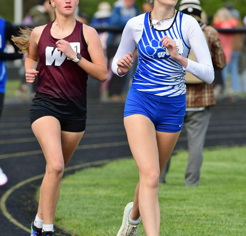 Two young female sprinters race on a curved track, one in a maroon top and black shorts, the other in a blue and white striped uniform.