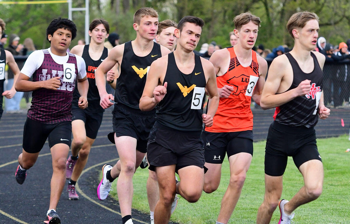 Group of male high school runners sprinting on a track during a race, bib numbers visible. Behind them, spectators watch from the sidelines.