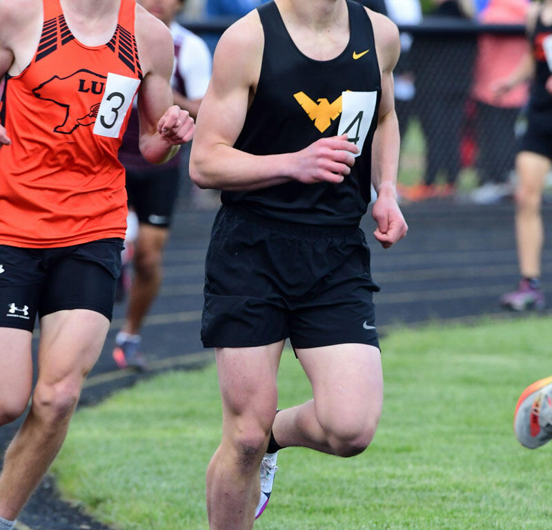 Two male athletes sprinting on a track during a race, wearing orange and black uniforms with numbers 3 and 4, spectators in the background.