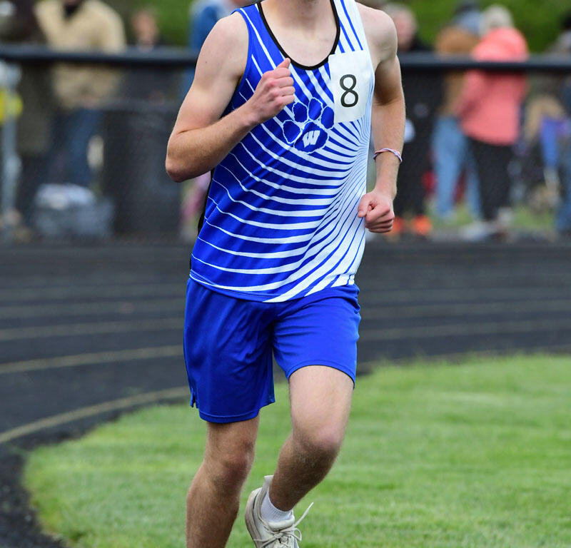 Young male athlete running on a track during a race, wearing a blue and white uniform with bib number 8, spectators in the blurred background.