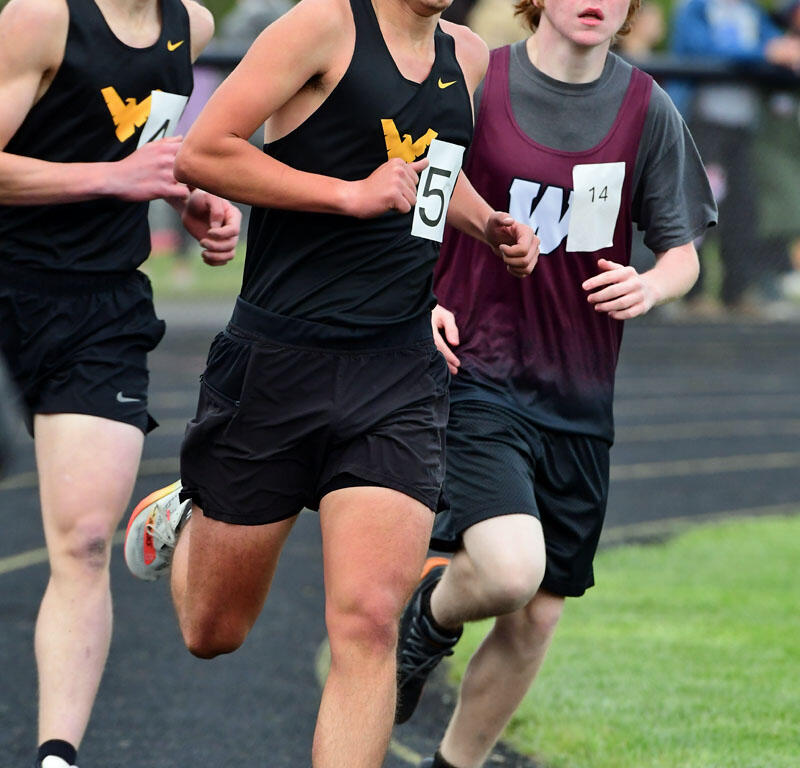 Three male high school runners in a sprint, wearing black and maroon jerseys on a track, competing closely.