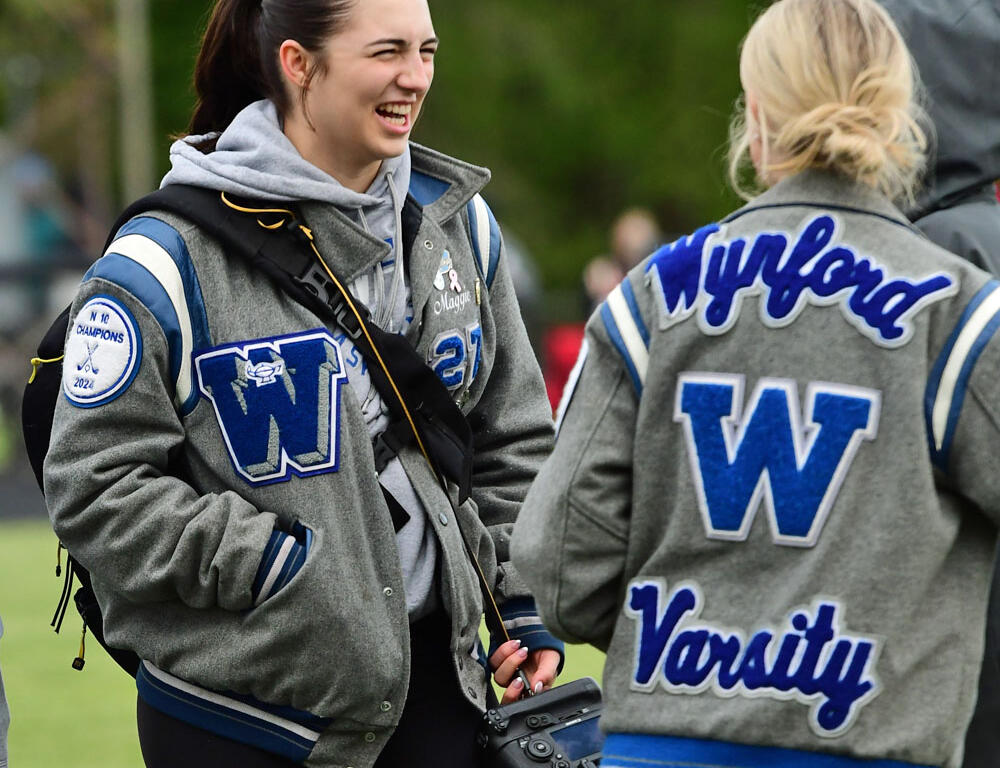 Two young women in gray varsity jackets chat and laugh on a field, one holding a camera strap across her chest.
