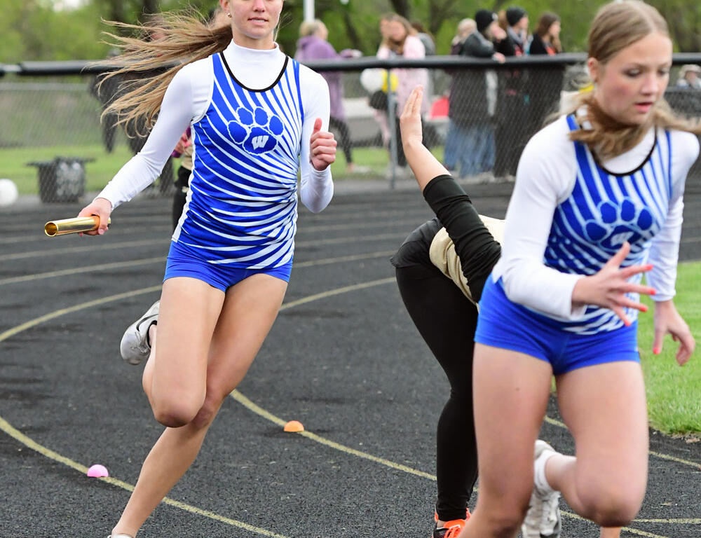 Two female sprinters run a relay on a track, one handing off a baton while wearing blue and white uniforms with paw print logos.