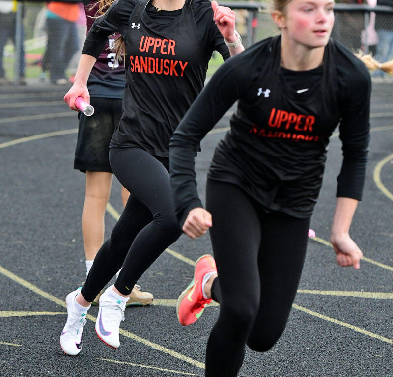 Female athletes in black Upper Sandusky uniforms sprint on an outdoor track during a race.