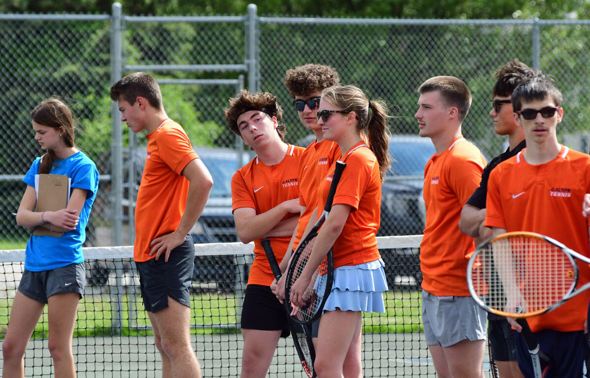 Group of tennis players standing along the net during practice, most in orange shirts, a girl in blue holding a clipboard nearby