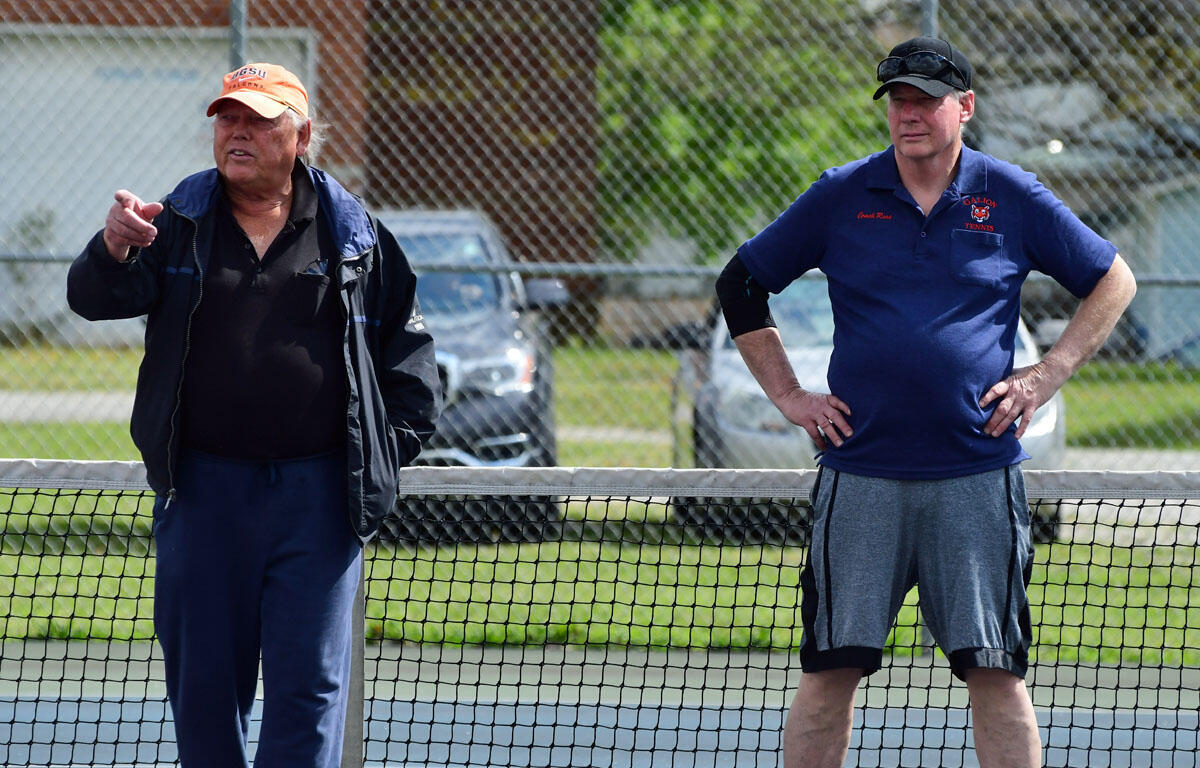 Two men standing at a tennis net outdoors; the man on the left points as the other stands with hands on hips, watching.