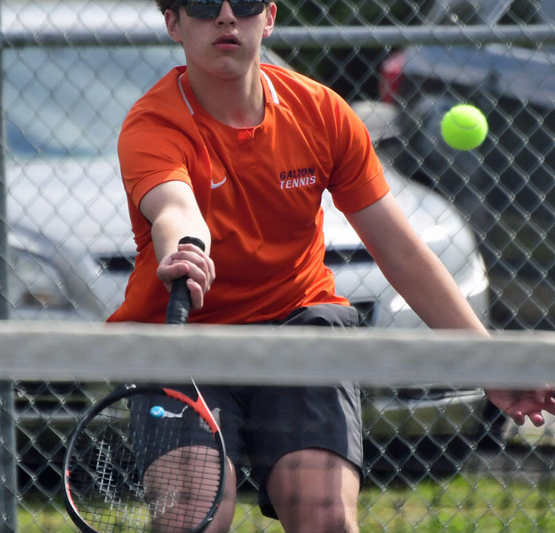 Young tennis player in an orange shirt and sunglasses reaches to hit a neon tennis ball with a racket on an outdoor court behind a chain-link fence.