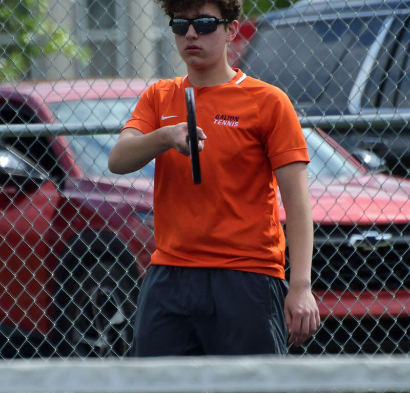 Young tennis player in an orange shirt and sunglasses holds a racket mid-swing as a tennis ball approaches on an outdoor court behind a chain-link fence.