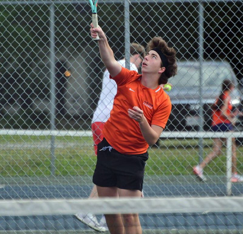 Tennis player in an orange shirt serves, racket raised high, on a fenced outdoor court.