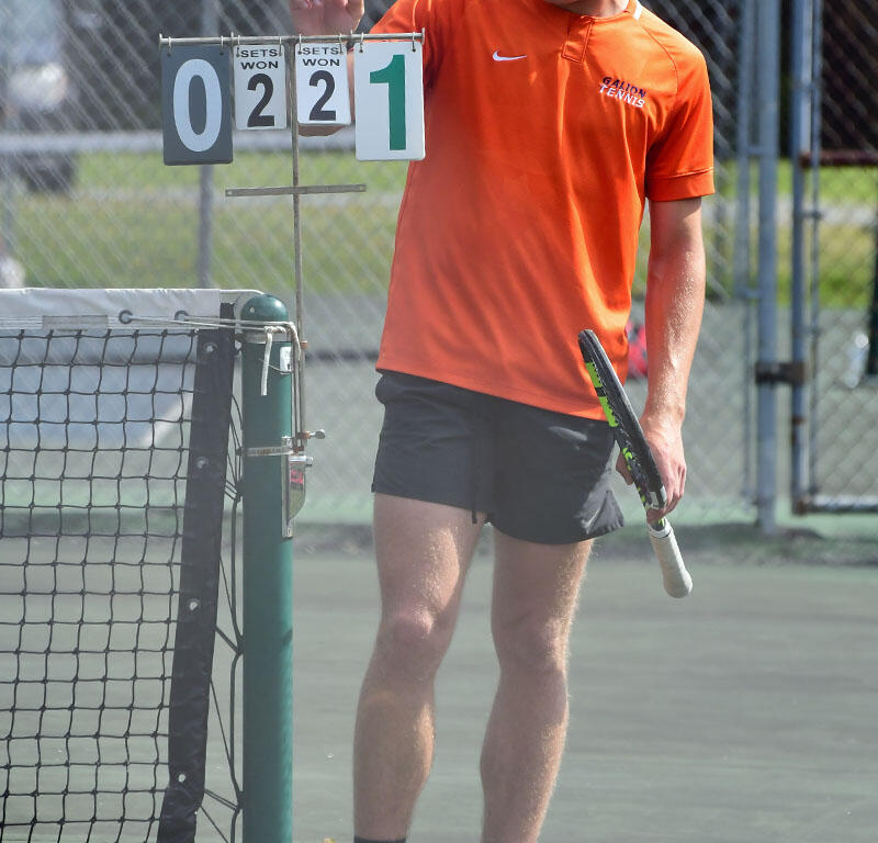 Male tennis player in an orange shirt adjusts a scoreboard on a tennis court, looking at the score display showing 0-2-2-1