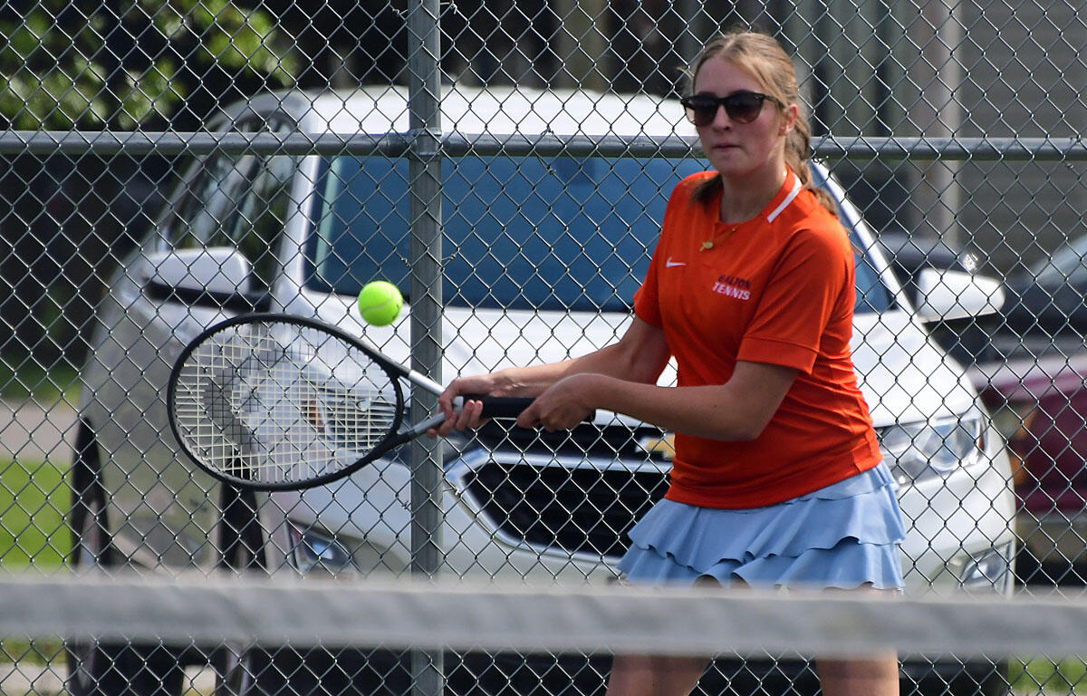 Female tennis player in an orange shirt and blue skirt swings her racket as a tennis ball approaches on a sunny outdoor court behind a chain-link fence.