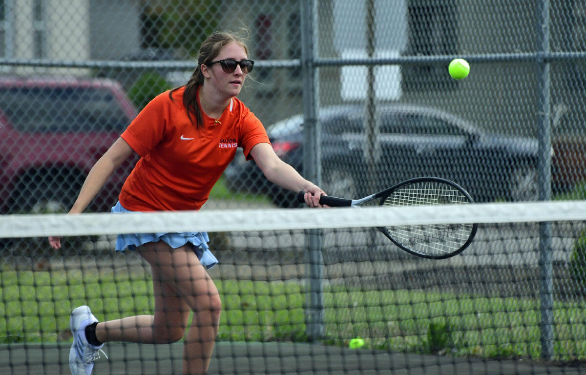 Female tennis player in an orange shirt and sunglasses reaches to hit a fast ball on an outdoor court behind a chain-link fence.
