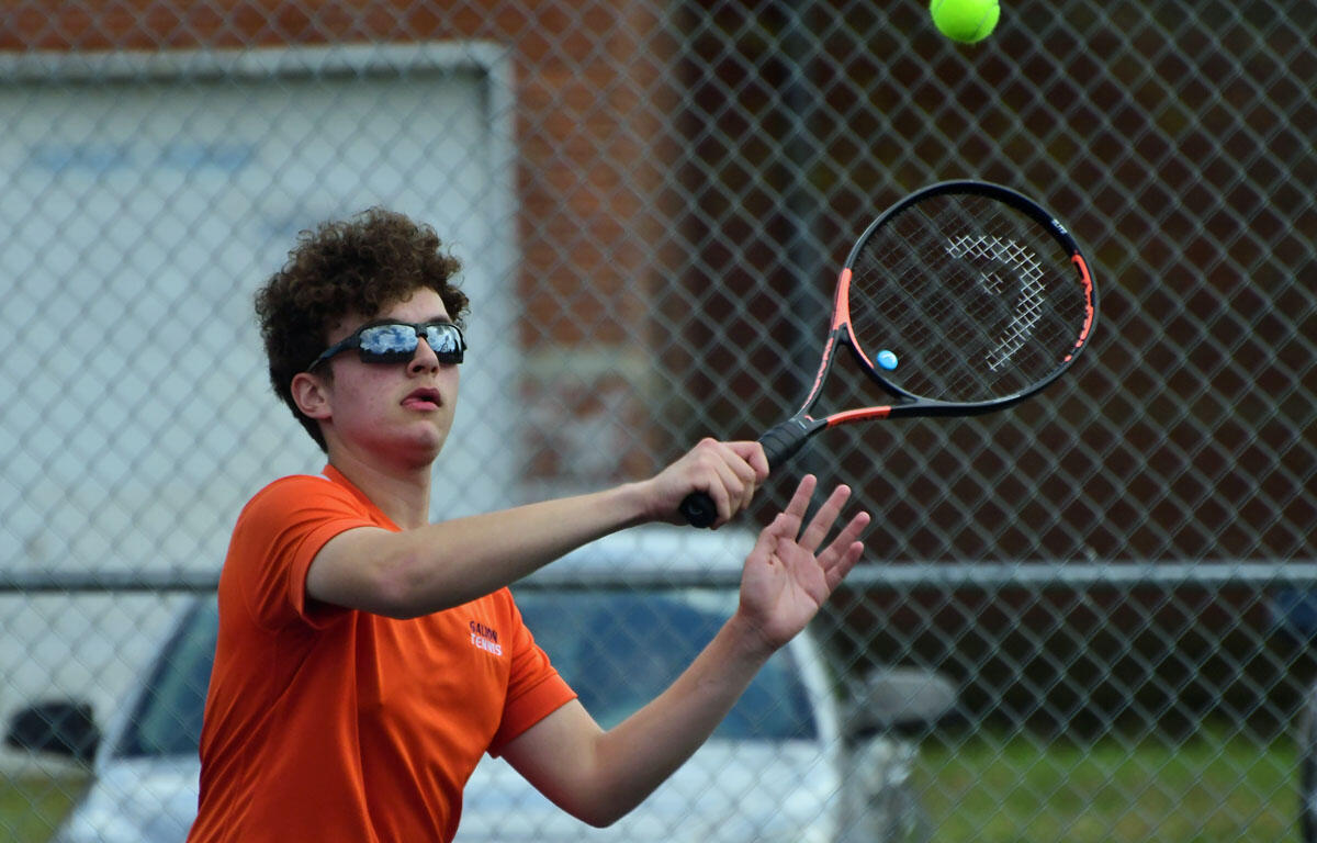 Young tennis player in an orange shirt and sunglasses reaches to hit a ball with a racket on a chain-link fenced court outdoors.