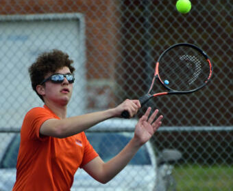 Young tennis player in an orange shirt and sunglasses reaches to hit a ball with a racket on a chain-link fenced court outdoors.