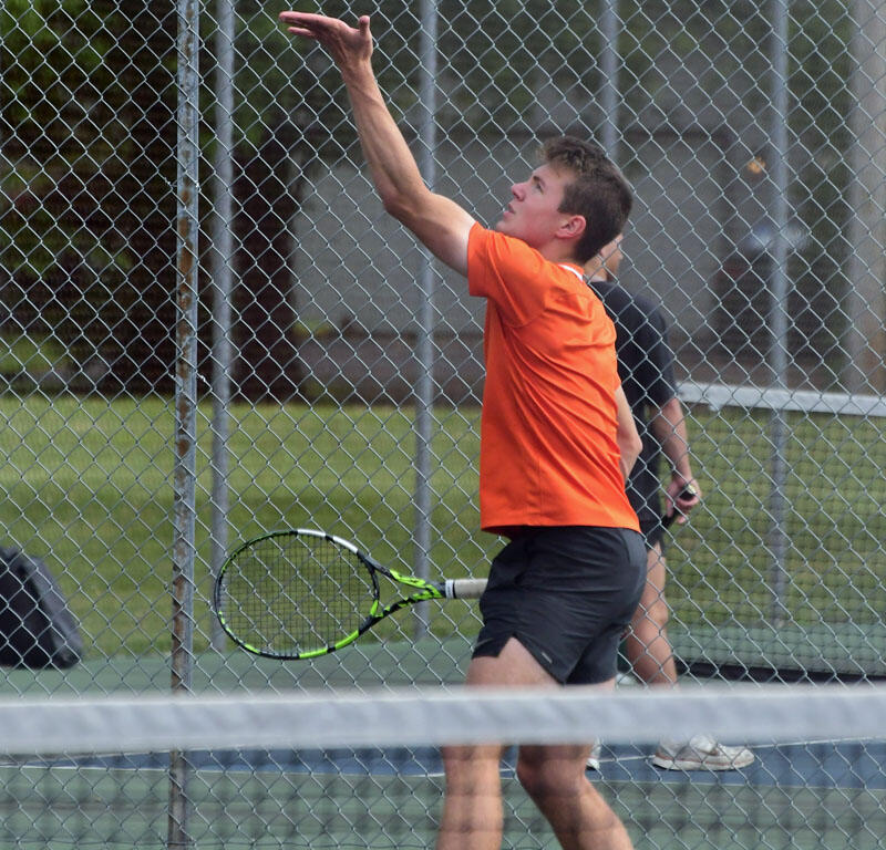 Male tennis player in an orange shirt serving on an outdoor court, ball in the air and racket at his side beside a chain-link fence.