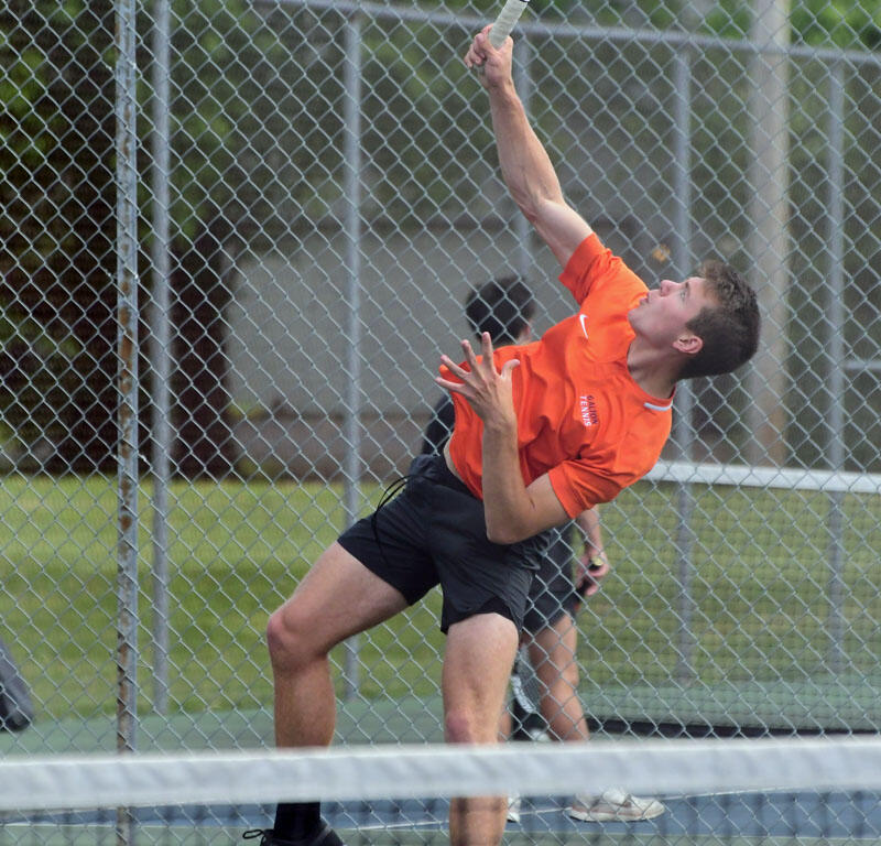 Tennis player in an orange shirt serves the ball on an outdoor court, mid-air with racket raised above head against a chain-link fence.
