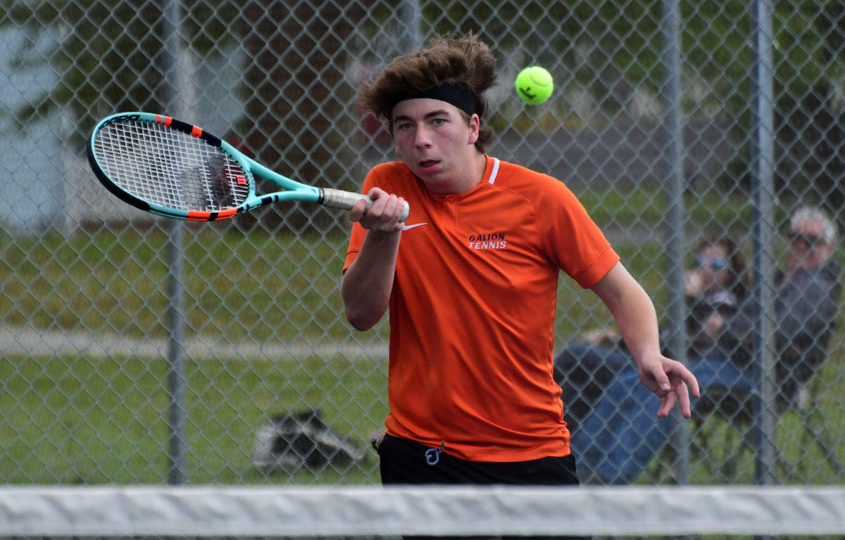 Tennis player in an orange Galion Tennis shirt hits a forehand volley on a fenced outdoor court, a bright tennis ball in midair nearby.