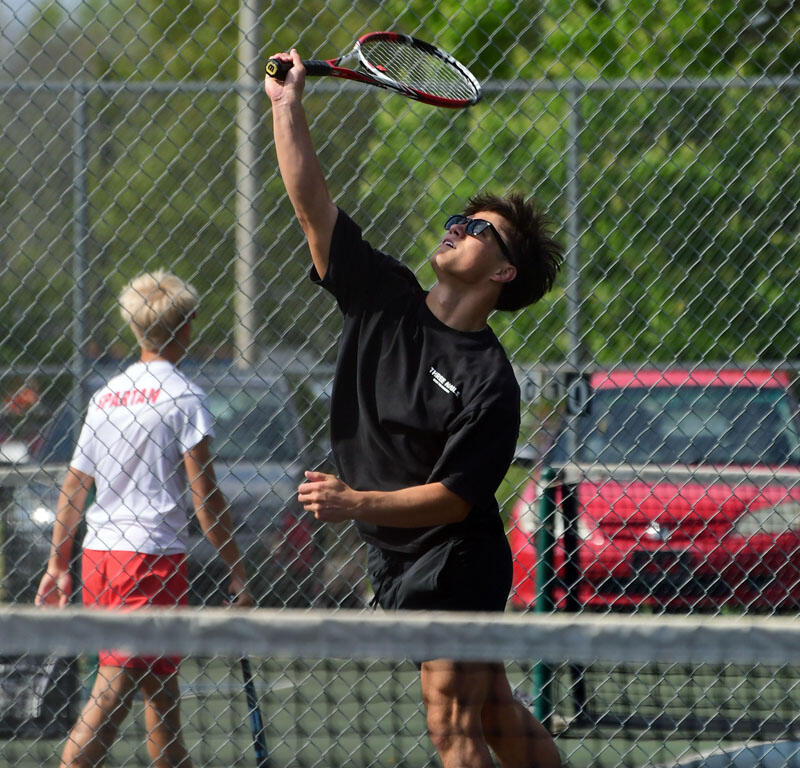 Tennis player in a black shirt serving the ball on an outdoor court, net in the foreground.