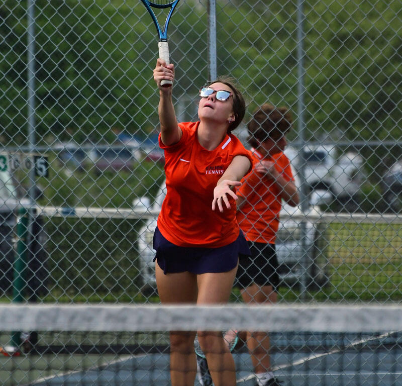 Female tennis player in an orange shirt leaps to hit a tennis ball with a blue racket on a chain-link fenced court.