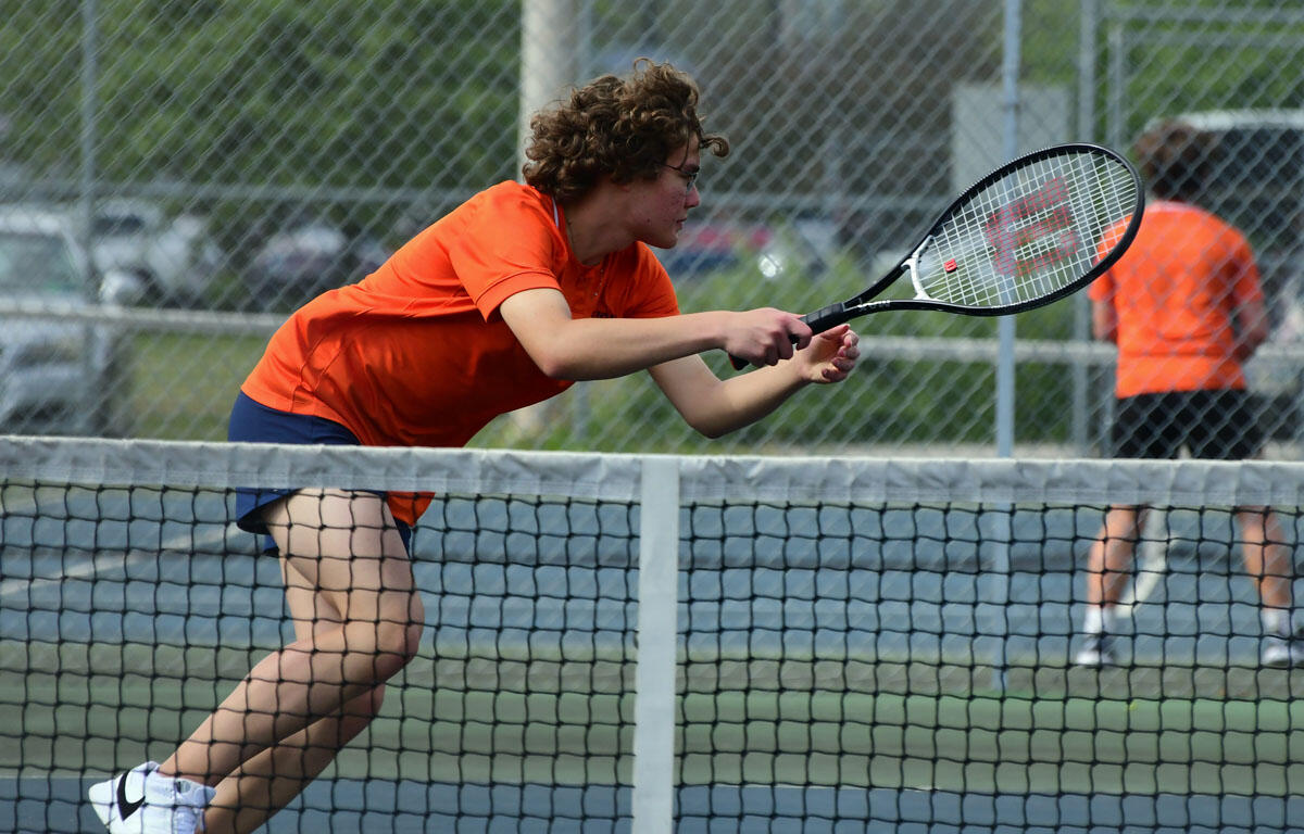 Tennis player in an orange shirt lunges forward with racket extended to hit a shot on an outdoor court, net in foreground and another player in orange in the background.