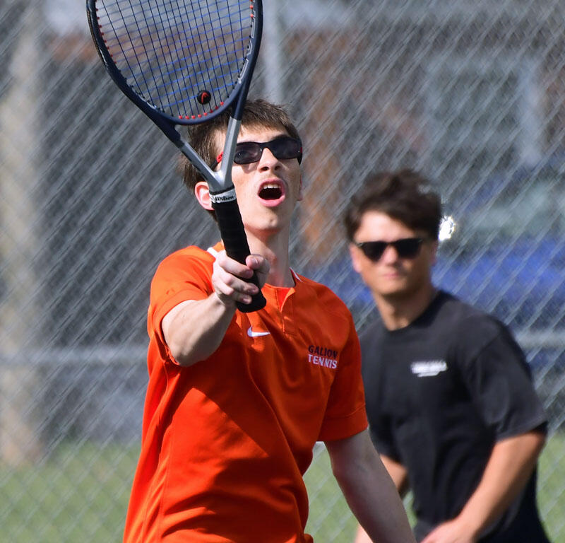 Male tennis player in orange shirt and sunglasses reaching to hit a forehand as a bright ball flies toward him near the net.