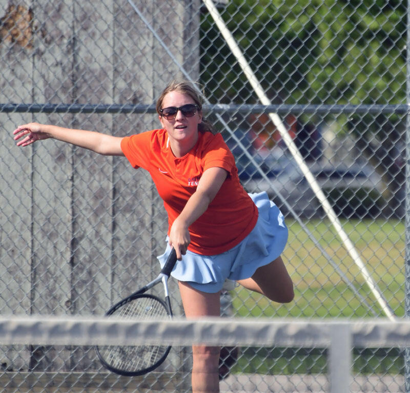 Female tennis player in an orange shirt and light-blue skirt serving on a court, ball in midair above her racket.