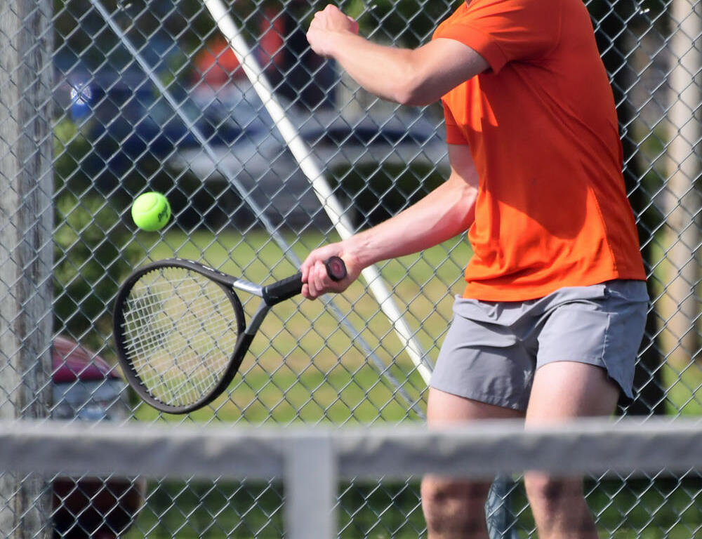 Tennis player in an orange shirt and gray shorts hits a ball on an outdoor court with a chain-link fence behind him.