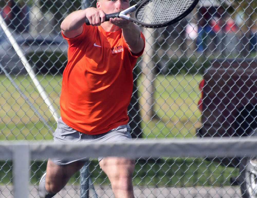 Male tennis player in an orange shirt hitting a two-handed backhand on a fenced outdoor court as a tennis ball flies toward him