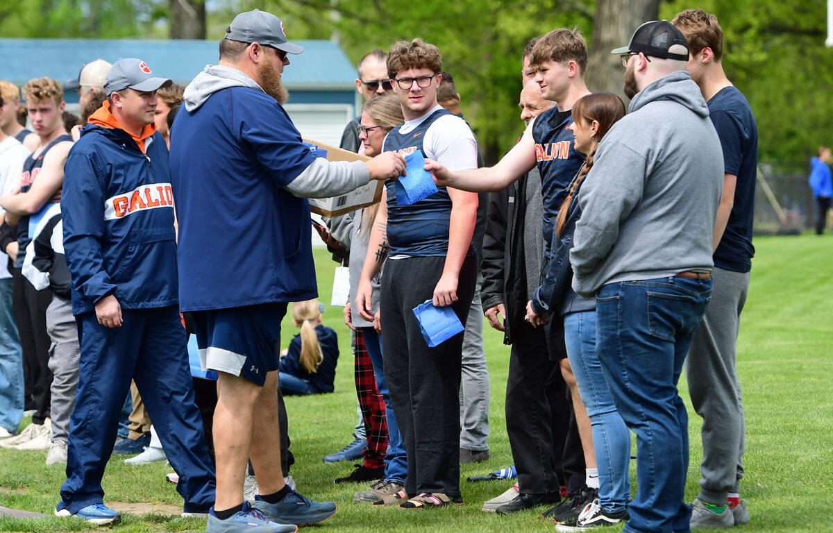 Coach in a blue jacket hands out blue award envelopes to a line of athletes on a grassy field during a team ceremony.