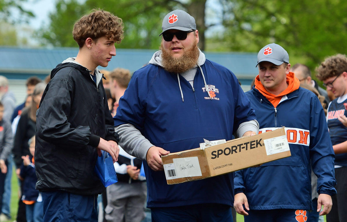 Bearded man in a blue jacket hands a SPORT-TEK box to a young man at an outdoor team event.